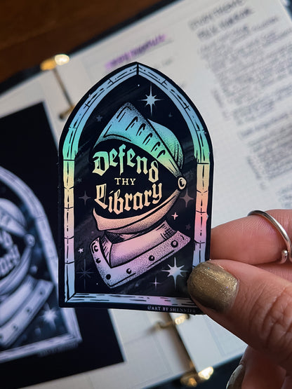 Holographic sticker with 'Defend thy Library' design held by a hand, with a blurred background.